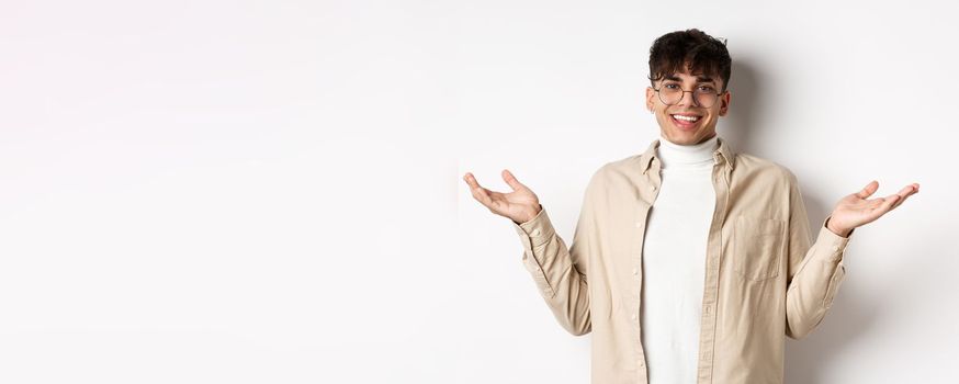 Portrait of happy and pleased young man spread hands sideways and smiling friendly, looking satisfied, standing in glasses on white background.