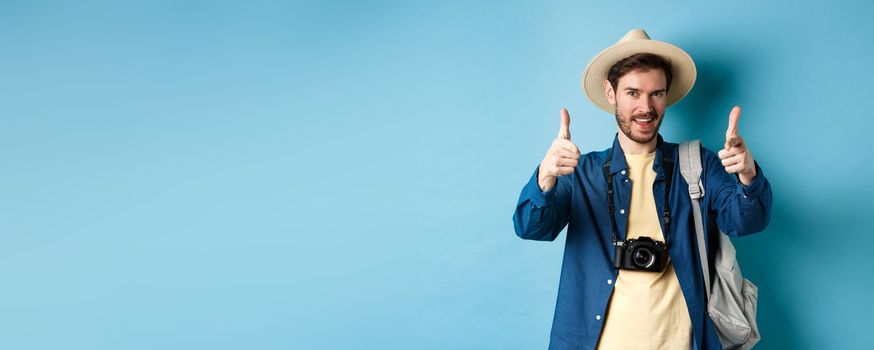 Handsome tourist going on summer vacation with camera and backpack pointing fingers at camera and inviting you, standing on blue background.