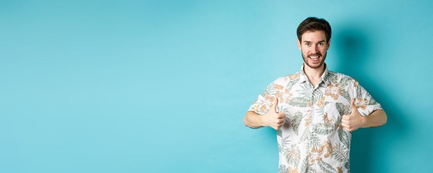 Handsome tourist showing thumbs up and say yes, praising good tourism agency, wearing hawaiian shirt, standing on blue background.