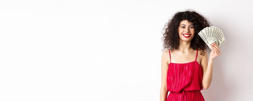 Stylish smiling woman in red dress showing money, standing on white background.