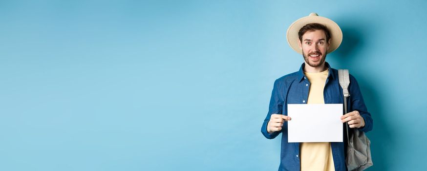 Cheerful tourist in summer hat, showing empty piece of paper and smiling, hitchhiking with backpack, standing on blue background.