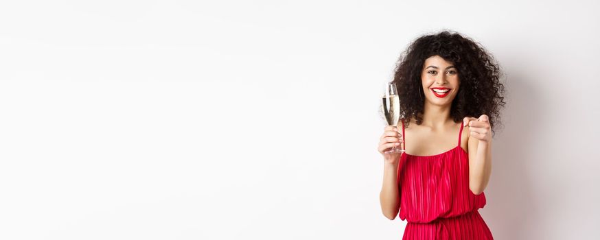 Beautiful curly woman in red dress, partying or having romantic date, holding glass of champagne and pointing at you, inviting person, standing on white background.