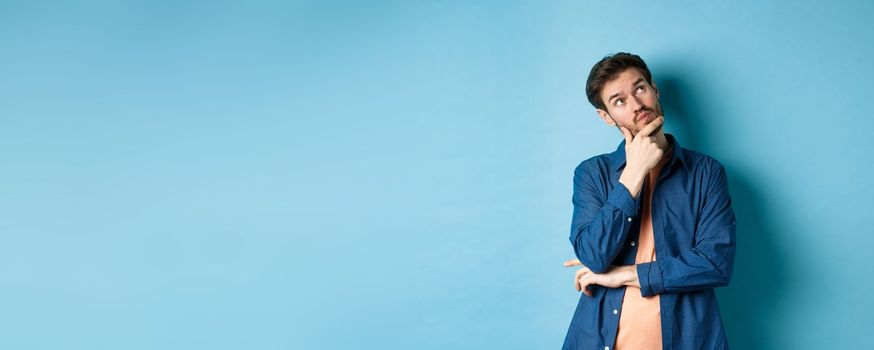 Pensive young man touching face and looking aside at empty space, imaging something, thinking off plan, standing on blue background.