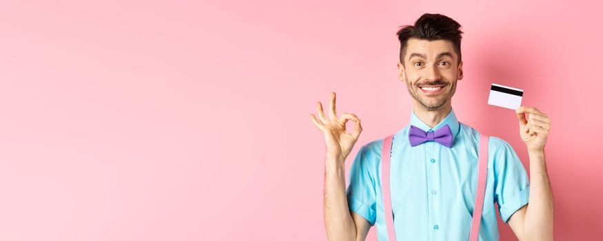 Shopping concept. Smiling handsome male shopper showing Ok sign and plastic credit card, buying something, standing satisfied on pink background.