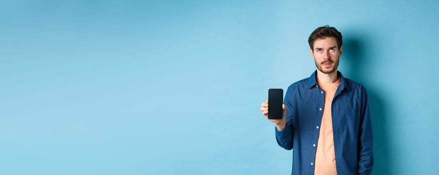 Annoyed young guy showing empty smartphone screen and roll eyes up bothered, standing on blue background.
