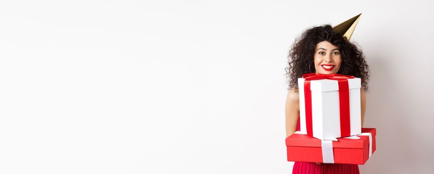 Holidays and celebration. Beautiful lady with curly hair, wearing party hat and holding big presents, smiling happy, receiving birthday gifts, standing on white background.