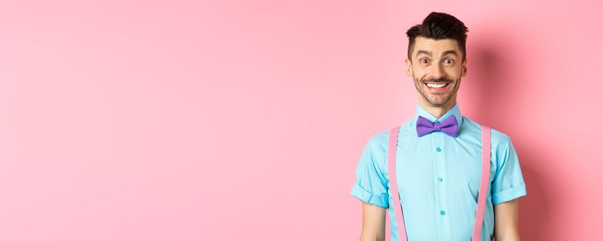 Happy young man with moustache smiling at camera, looking excited and cheerful, standing on pink background in bow-tie and shirt.