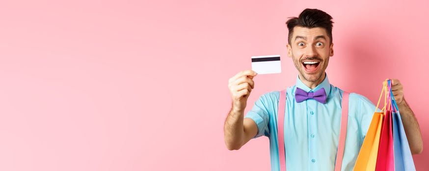 Happy young man giving his credit card and holding shopping bags, buying with discounts, standing on pink background and smiling.