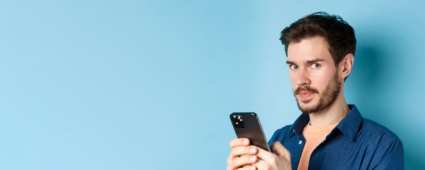 Close-up of handsome european guy using smartphone and looking at camera, standing on blue background.