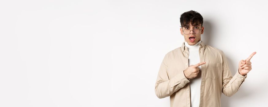 Shocked young man in glasses pointing fingers right at empty space, asking about advertisement, standing on white background.