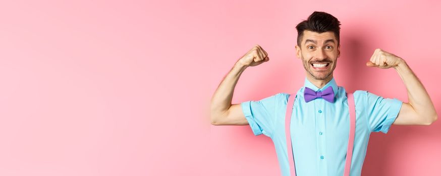 Smiling caucasian man with bow-tie and suspenders, showing muscles and feeling strong, flexing biceps to show-off, standing over pink background.