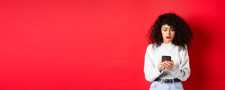 Sad and worried woman reading message on smartphone, receive bad news on social media, standing against red background.