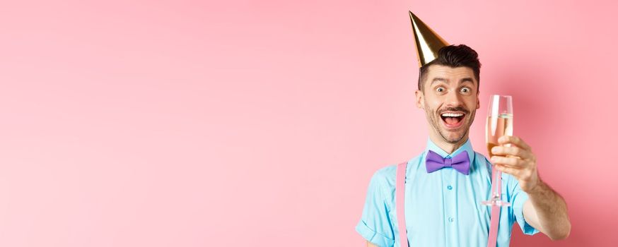 Holidays and celebration concept. Excited birthday guy raising glass of champagne, making toast or speech, standing in party hat and having fun on pink background.