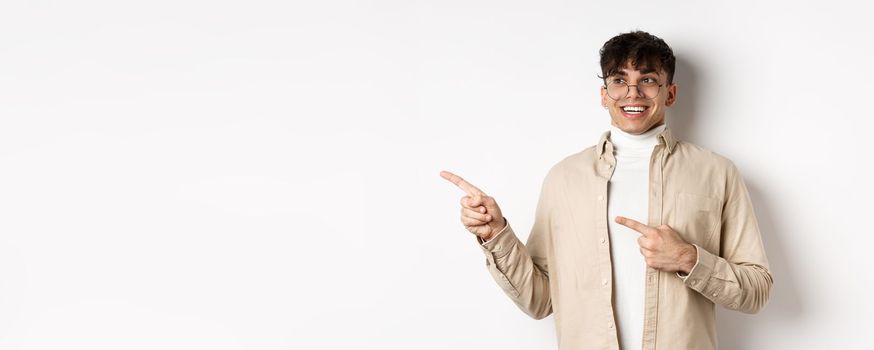 Excited and enthusiastic young man in glasses, pointing fingers and looking left with happy smile, standing in awe on white background.