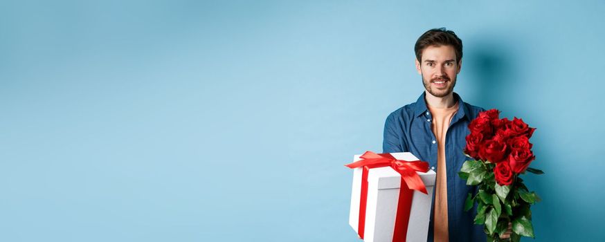 Love and Valentines day concept. Charming young man giving gift and bouquet of roses to girlfriend, standing over blue background.