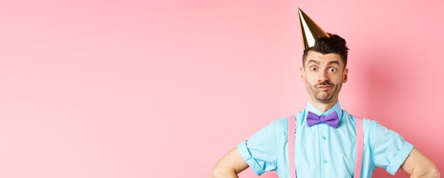 Holidays and celebration concept. Close-up of confused male entertainer in party hat and bow-tie, looking puzzled and shocked, standing over pink background.