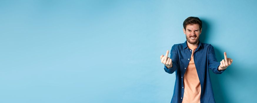 Angry caucasian man say fuck you and showing middle fingers, make rude gesture with pissed-off face, standing annoyed on blue background.