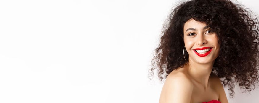 Close-up of cheerful smiling woman with red lipstick and curly hairstyle, looking happy, standing over white background.