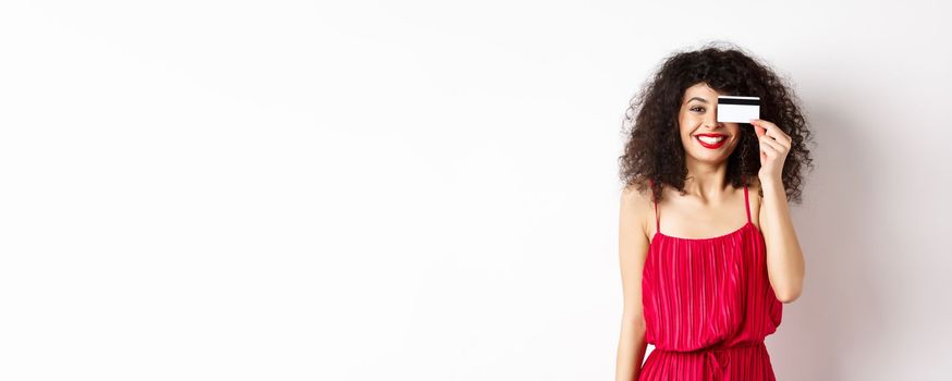 Image of elegant woman in red dress, showing plastic credit card and smiling at camera, standing over white background. Copy space