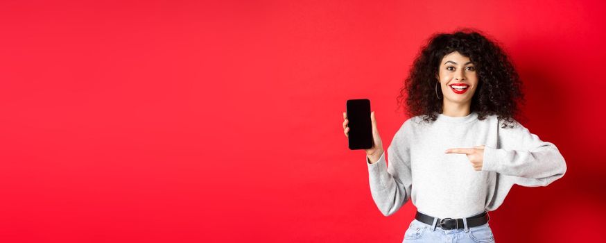 Beautiful female model showing empty smartphone screen, pointing at phone display and smiling, standing against red background.