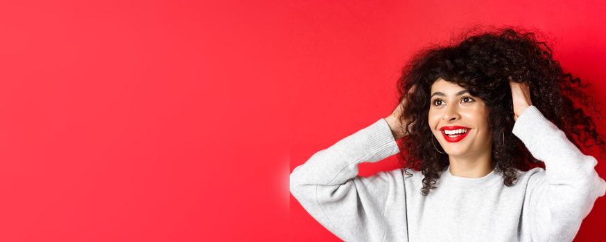 Happy beautiful woman touching natural curly hair and smiling pleased, looking left, like her hairstyle, standing against red background.