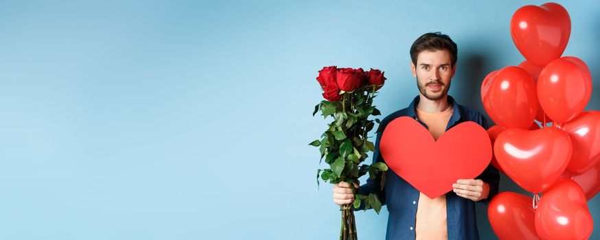 Valentines day romance. Young man with bouquet of red roses and heart balloons smiling, bring presents for lover on valentine date, standing over blue background.