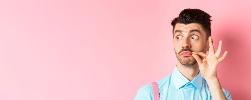 Close up of young caucasian man touching moustache and pucker lips, staring left at logo, standing on pink background.