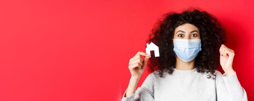 Real estate and covid-19 concept. Excited woman in medical mask showing small paper house cutout, standing on red background.