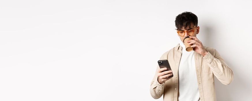 Real people. Young man reading smartphone screen and drinking coffee, looking at phone, standing on white background.