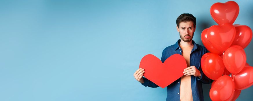 Sad man looking heartbroken and lonely, holding paper red heart and standing near balloons over blue background.
