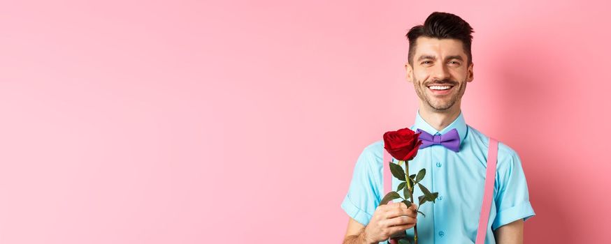 Attractive fancy guy waiting for date on lovers date, holding red rose and smiling, standing on romantic pink background. Valentines day concept.