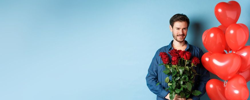 Romantic boyfriend winking and smiling, holding bouquet of flowers on Valentines day, standing near heart balloons for lover, blue background.