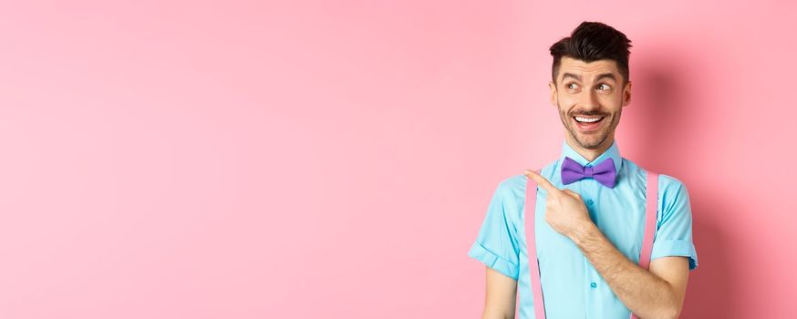 Cheerful caucasian man in funny bowtie looking and pointing hand left, showing promo logo, standing on pink background. Copy space