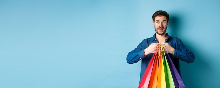 Happy young man holding colorful shopping bags and smiling excited, buying gifts, standing on blue background.