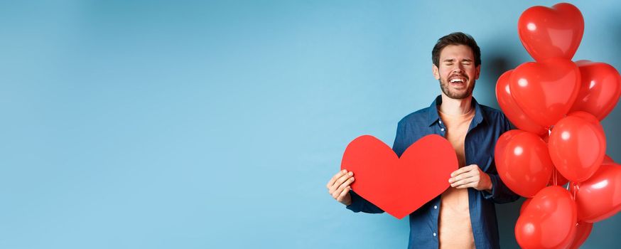 Heartbroken man crying of breakup of valentines day, holding red heart cutout and standing near romantic balloons over blue background.