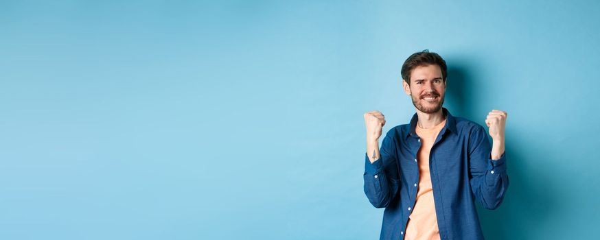 Image of happy man celebrating success, shouting yes and shaking clenched fists, achieve goal and triumphing, winning and dancing, standing on blue background.