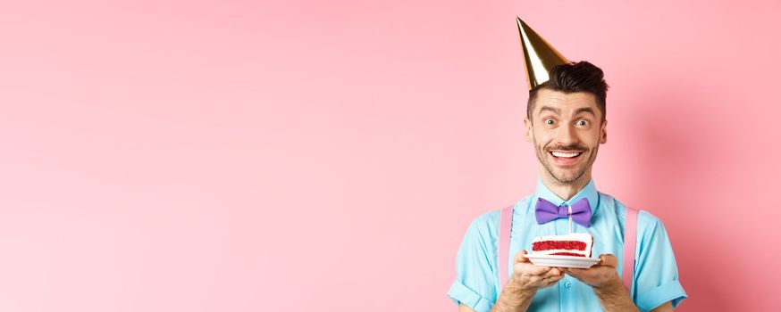 Holidays and celebration concept. Cheerful young man celebrating birthday in party hat, holding b-day cake with candle and making wish, smiling happy at camera, pink background.
