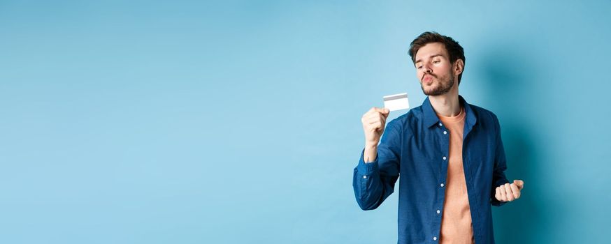 Funny guy kissing his plastic credit card and looking satisfied, shopping with discounts, standing on blue background.