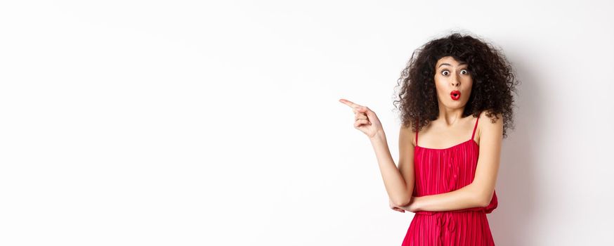 Surprised elegant woman in red dress, saying wow and pointing finger left, showing interesting promo, standing over white background.