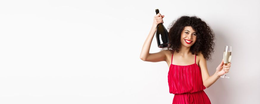 Happy woman partying on valentines day holiday, dancing with glass and bottle of champagne, wearing red dress, smiling on white background.