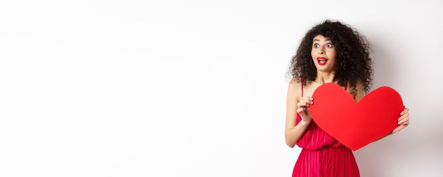 Valentines day. Young woman looking surprised left, seeing something amazing on lovers day, found true love, holding big red heart on chest, white background.