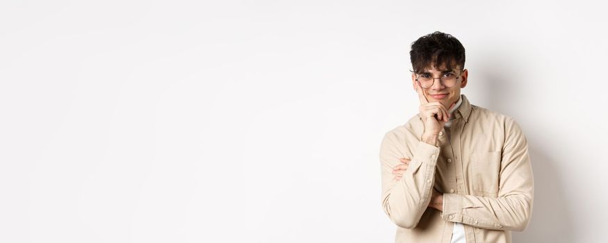 Cunning young man watching something interesting happen, lean face on arm and smiling devious, looking at camera intrigued, standing on white background.