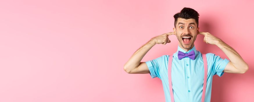 Childish funny guy shut ears and laughing at camera, refuse to listen, standing ignorant on pink background.