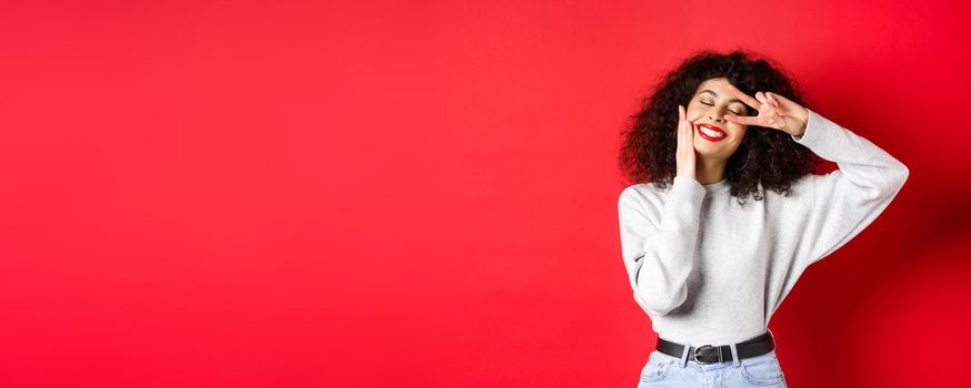 Beauty. Pretty lady with curly hair and red lips, touching face with makeup and showing v-sign on eye, smiling carefree, standing against studio background.