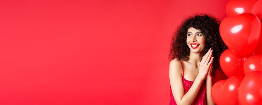 Excited beautiful woman with curly hair, standing near heart balloons and rubbing palms together, expect good deal or relish, red background.