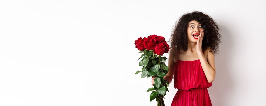 Excited curly woman in red dress, receive bouquet of roses and look surprise, rejoicing from romantic gift, standing on white background.