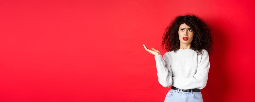 Annoyed and puzzled young woman with curly hair, raising hands up and looking aside, cant understand something strange, standing on red background.