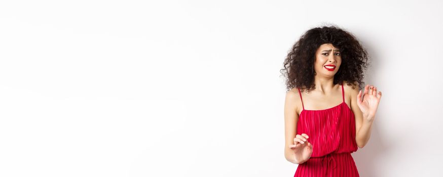 Young woman cringe of something creepy, grimacing and recoil from disgusting awful thing, standing in red dress on white background.