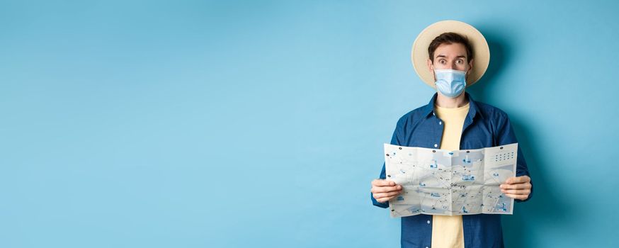 Covid-19, pandemic and travel concept. Tourist in medical mask look surprised, holding map, standing on blue background.