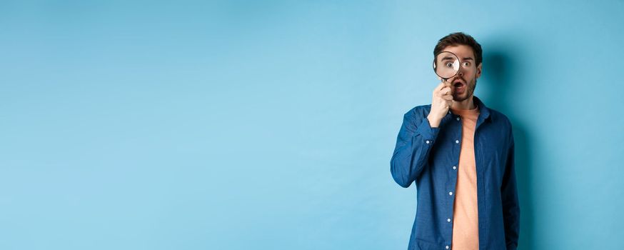 Impressed man look in awe through magnifying glass, drop jaw and stare at camera, standing on blue background.